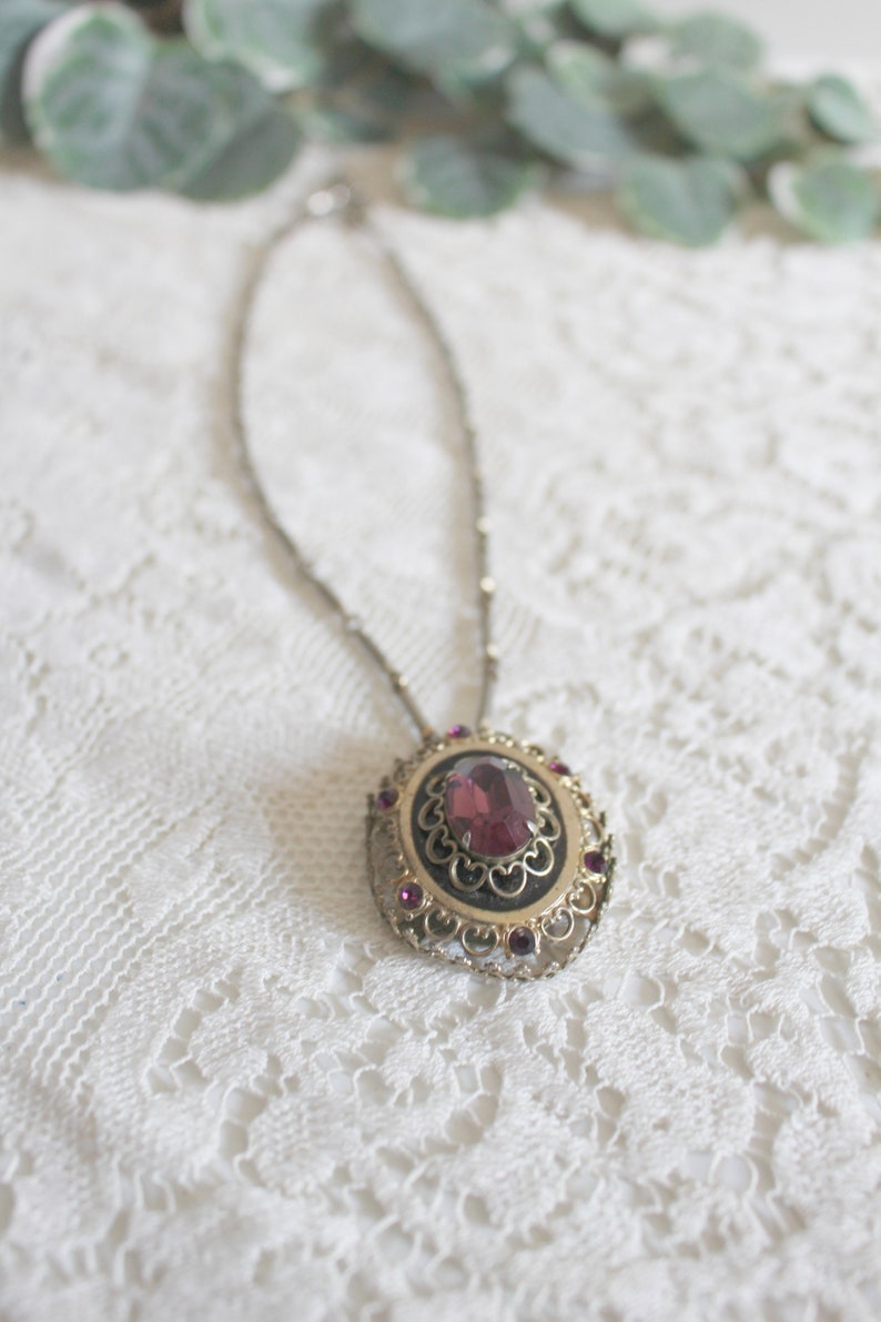 May include: A silver chain necklace with a pendant featuring a large, oval-shaped, purple gemstone surrounded by a gold filigree design and smaller purple gemstones.