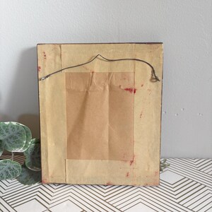 Puede incluir: Parte trasera de una obra de arte enmarcada rectangular. El marco marr&oacute;n oscuro tiene un papel de respaldo marr&oacute;n claro. Se adjunta un cable negro para colgar. El papel de respaldo tiene un trozo rectangular de papel marr&oacute;n adherido. Manchas rojas visibles.