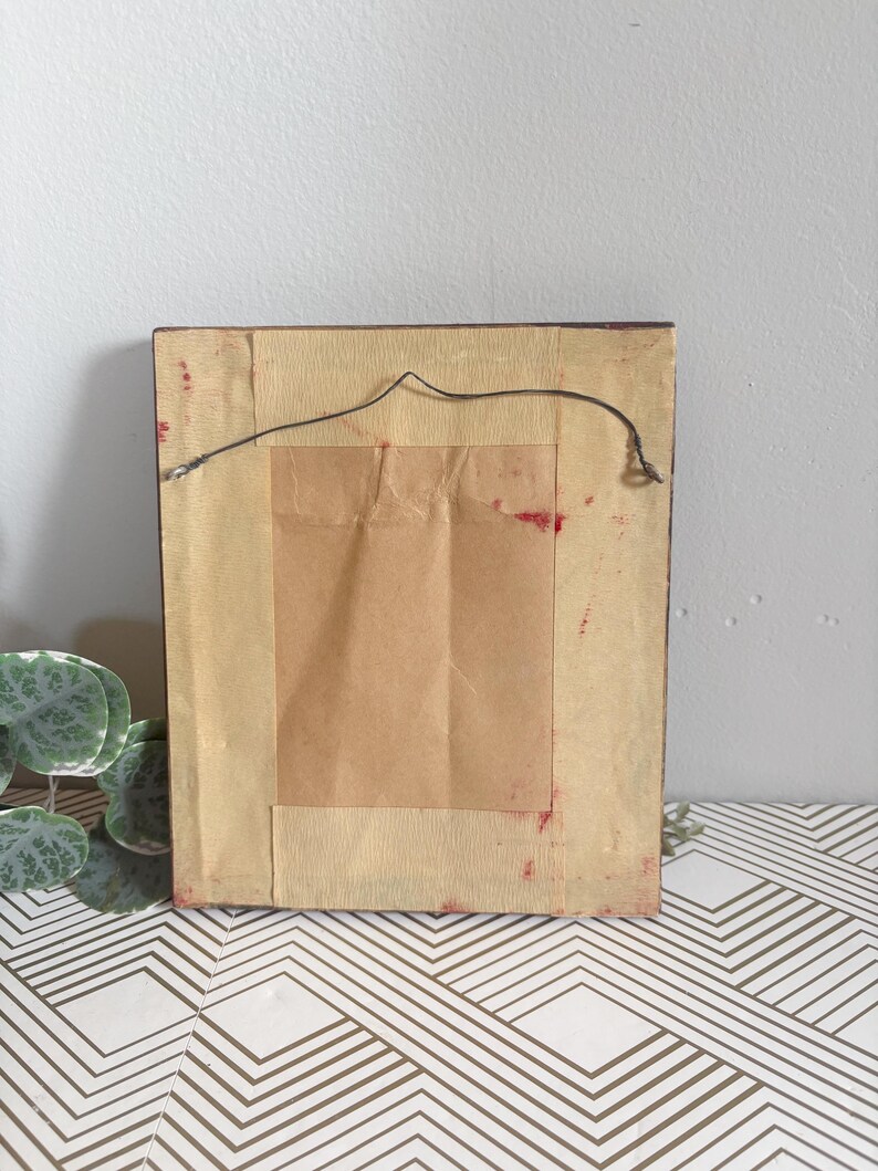 Puede incluir: Parte trasera de una obra de arte enmarcada rectangular. El marco marr&oacute;n oscuro tiene un papel de respaldo marr&oacute;n claro. Se adjunta un cable negro para colgar. El papel de respaldo tiene un trozo rectangular de papel marr&oacute;n adherido. Manchas rojas visibles.