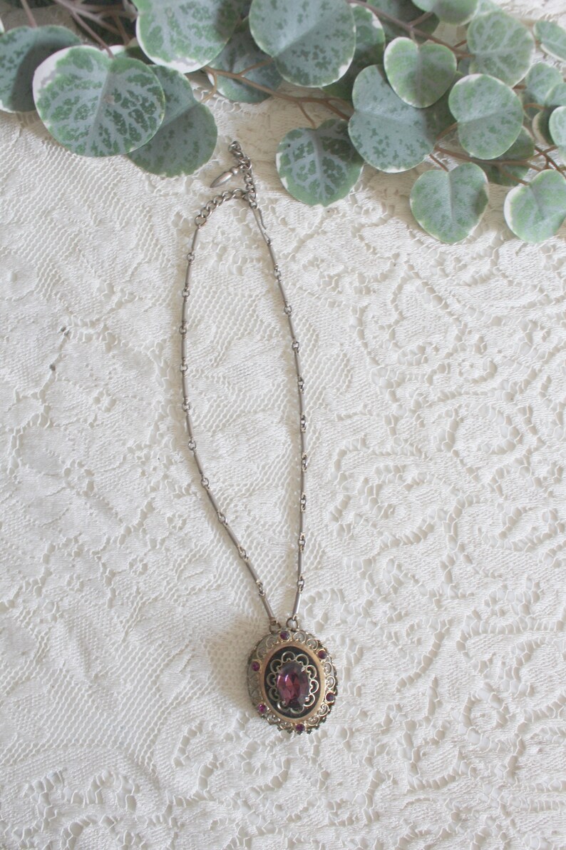 May include: A silver chain necklace with a round pendant featuring a large purple gemstone surrounded by smaller purple gemstones and a gold filigree design.