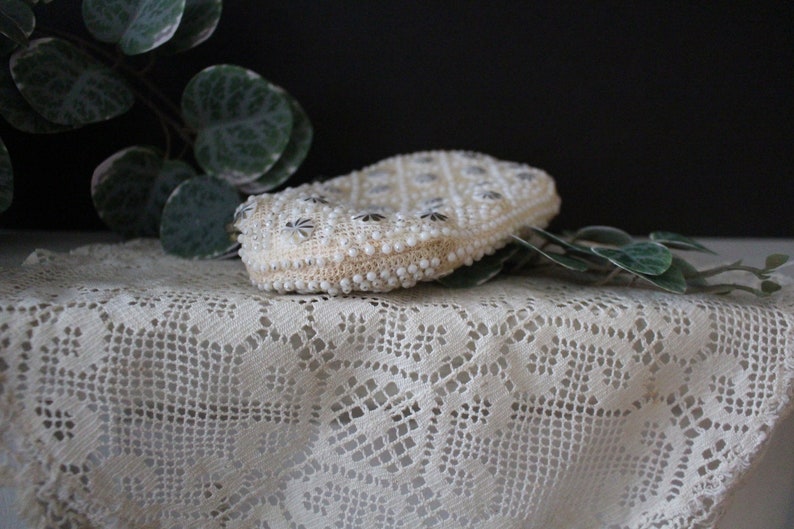 May include: A white beaded clutch purse with a silver clasp. The purse is decorated with a repeating pattern of small silver stars.