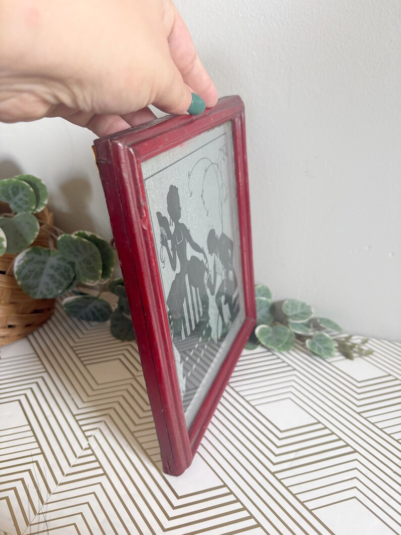 Puede incluir: Una obra de arte de silueta enmarcada vintage. El marco es de color rojo desgastado. La obra de arte representa una silueta negra de figuras, posiblemente una familia, sobre un fondo gris claro. El marco se sostiene en &aacute;ngulo, mostrando la obra de arte.