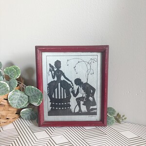 Puede incluir: Arte de silueta enmarcado que presenta una imagen en blanco y negro de un hombre arrodillado ante una mujer. La obra de arte est&aacute; enmarcada en un marco de madera rojo. El fondo es de color gris claro.