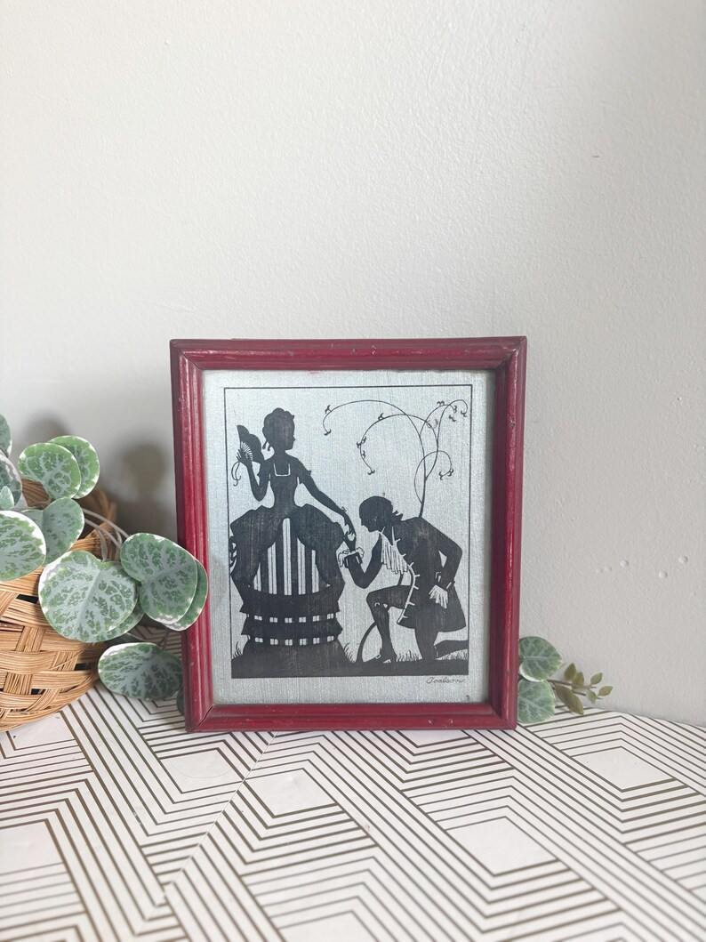 Puede incluir: Arte de silueta enmarcado que presenta una imagen en blanco y negro de un hombre arrodillado ante una mujer. La obra de arte est&aacute; enmarcada en un marco de madera rojo. El fondo es de color gris claro.