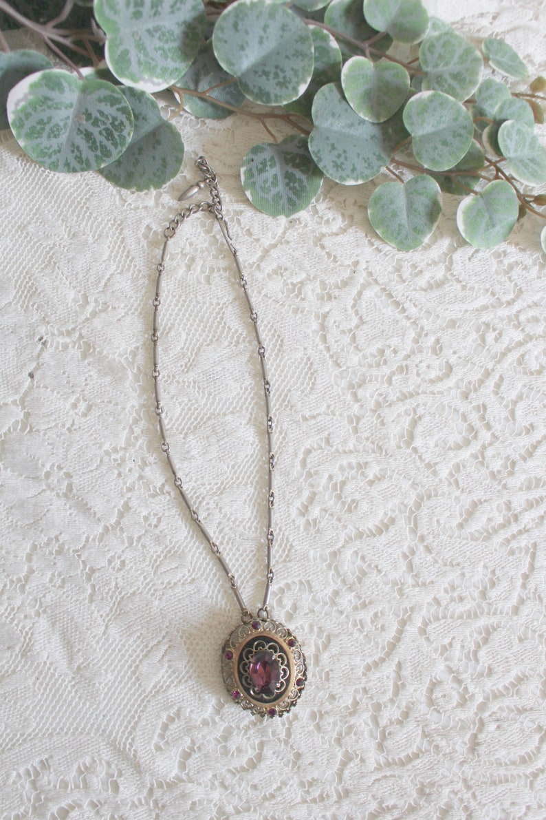 May include: A silver chain necklace with a pendant featuring a large, oval-shaped, purple gemstone surrounded by a gold filigree design and smaller purple gemstones.