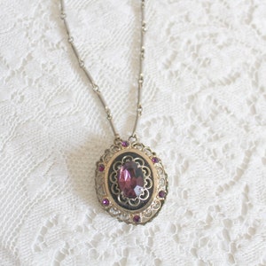 May include: A silver chain necklace with a pendant featuring a large, oval-shaped, dark purple gemstone surrounded by a filigree design and smaller purple gemstones.