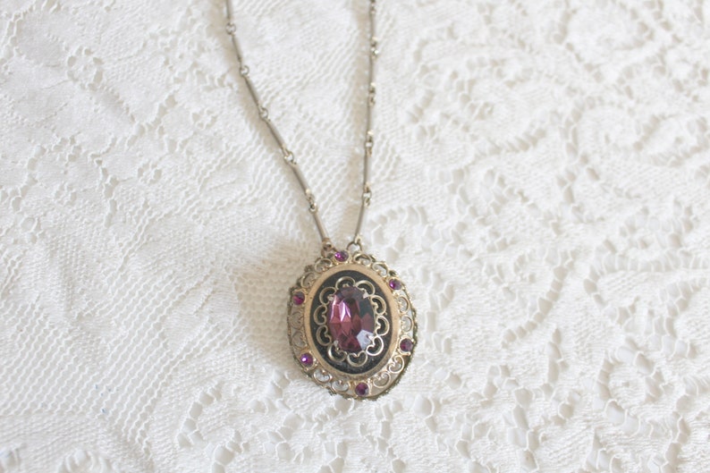 May include: A silver chain necklace with a pendant featuring a large, oval-shaped, dark purple gemstone surrounded by a filigree design and smaller purple gemstones.