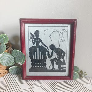 Puede incluir: Arte de silueta enmarcado que presenta una escena rom&aacute;ntica. La imagen en blanco y negro representa a una mujer sentada y a un hombre arrodillado, sobre un fondo claro. El marco es de color rojo oscuro, lo que a&ntilde;ade un toque vintage a la obra.