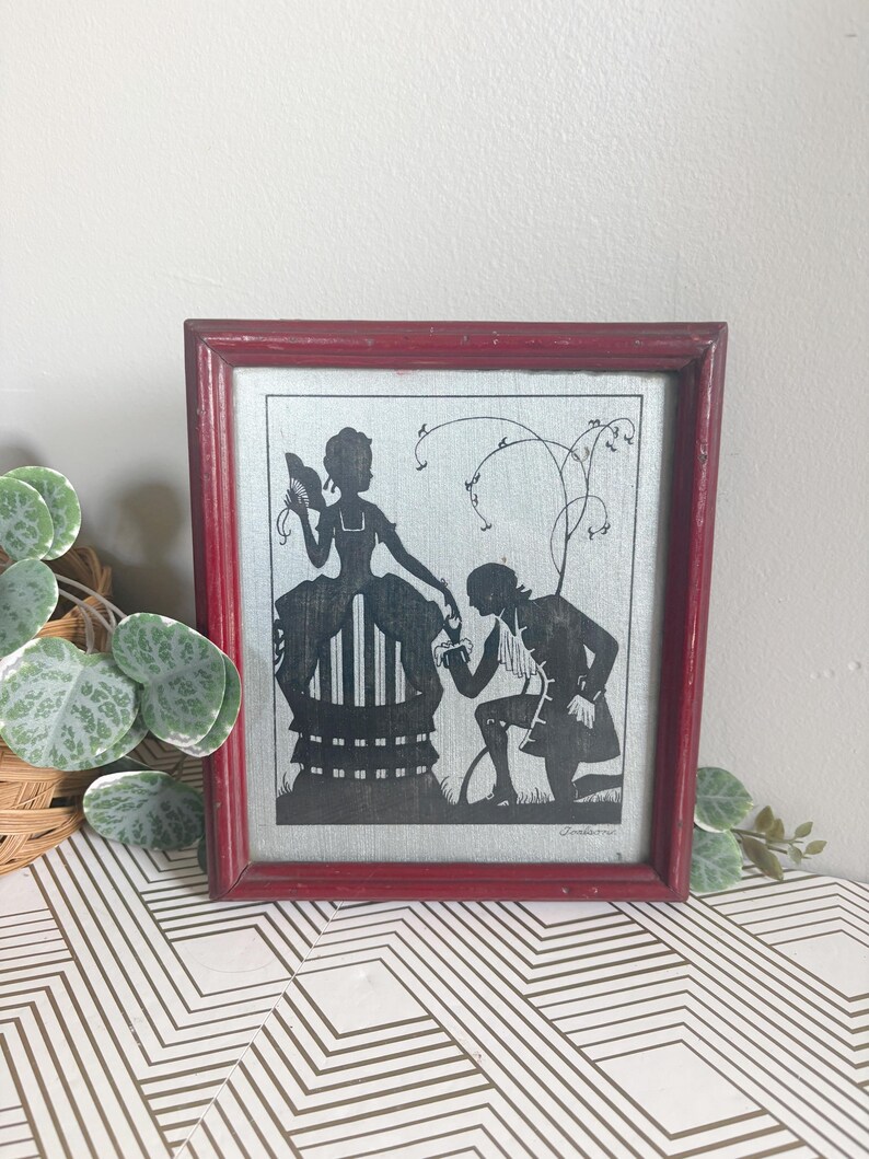 Puede incluir: Arte de silueta enmarcado que presenta una escena rom&aacute;ntica. La imagen en blanco y negro representa a una mujer sentada y a un hombre arrodillado, sobre un fondo claro. El marco es de color rojo oscuro, lo que a&ntilde;ade un toque vintage a la obra.