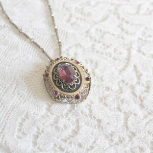 May include: A silver chain necklace with a pendant featuring a large, oval-shaped, purple gemstone surrounded by smaller, purple gemstones in a filigree setting.