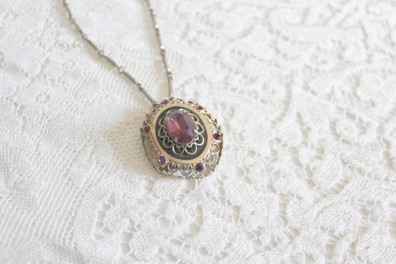 May include: A silver chain necklace with a pendant featuring a large, oval-shaped, purple gemstone surrounded by smaller, purple gemstones in a filigree setting.