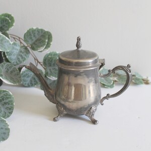 May include: A silver teapot with a lid and three feet. The teapot has a decorative handle and a small knob on the lid.