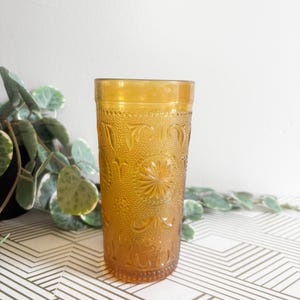 May include: Amber-coloured vintage drinking glass with an embossed floral and dot pattern. The tall glass has a textured surface and a wide rim. The glass is empty and stands on a white and gold geometric patterned surface.