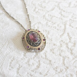 May include: A silver chain necklace with a pendant featuring a large, oval-shaped, purple gemstone surrounded by smaller, purple gemstones in a filigree setting.