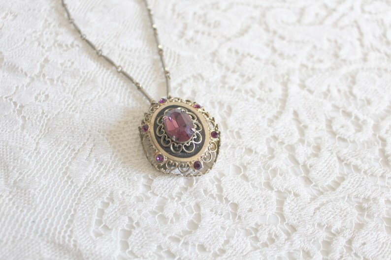 May include: A silver chain necklace with a pendant featuring a large, oval-shaped, purple gemstone surrounded by smaller, purple gemstones in a filigree setting.