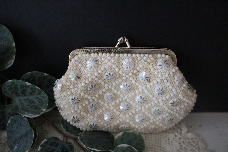 May include: A white beaded clutch purse with a silver floral design and a gold clasp.