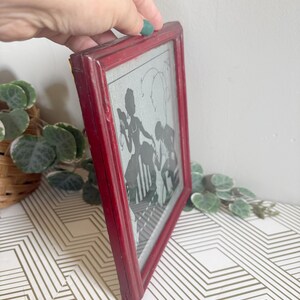 Puede incluir: Una obra de arte de silueta enmarcada vintage. El marco es de color rojo desgastado. La obra de arte en el interior presenta siluetas negras de figuras sobre un fondo gris claro. La obra de arte se exhibe sobre una superficie con un patr&oacute;n geom&eacute;trico blanco y dorado.