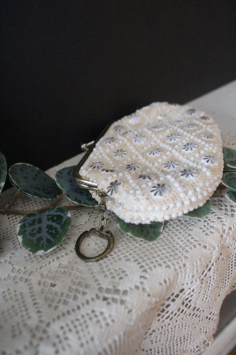 May include: A white beaded coin purse with a silver clasp and a key ring. The purse is on a white crocheted surface.