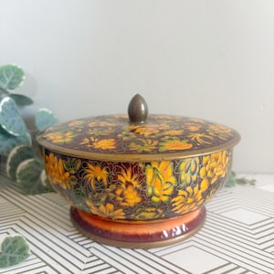 May include: A vintage, round decorative bowl with a lid. The bowl is adorned with a floral pattern in shades of yellow, orange, and green against a dark background. It sits on a matching pedestal base and has a small, brown knob on top.