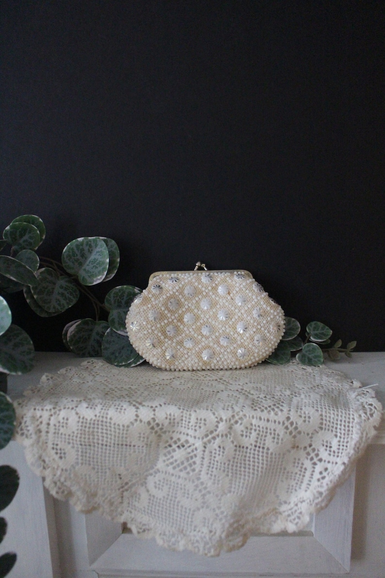 May include: A white beaded clutch purse with a silver clasp. The purse is sitting on a white crocheted doily.