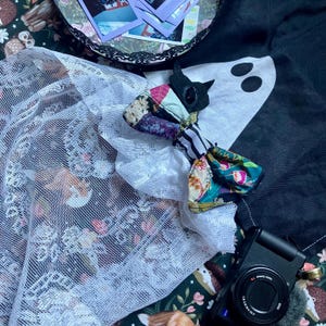 May include: A black cat-shaped plush toy featuring a patchwork fabric body with floral, striped, and solid patterns, accented by a white mesh fabric overlay. The toy is positioned near a black camera and a dark fabric with a white ghost graphic.