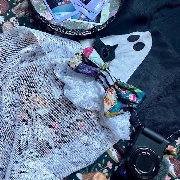 May include: A black cat-shaped plush toy featuring a patchwork fabric body with floral, striped, and solid patterns, accented by a white mesh fabric overlay. The toy is positioned near a black camera and a dark fabric with a white ghost graphic.