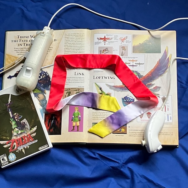 May include: A white video game controller with a cord, a video game case for The Legend of Zelda: Skyward Sword, and a long, rectangular fabric strip featuring a red, white, purple, and yellow gradient, all resting on an open illustrated book.