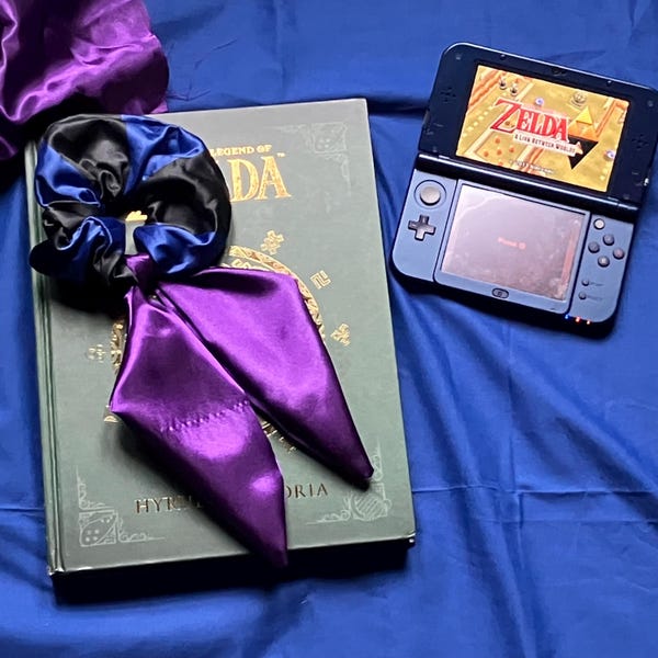 May include: A hair scrunchie featuring a black and blue satin fabric band with two long, pointed purple satin tails. The scrunchie rests on a green hardcover book titled The Legend of Zelda: Hyrule Historia next to a black handheld game console.