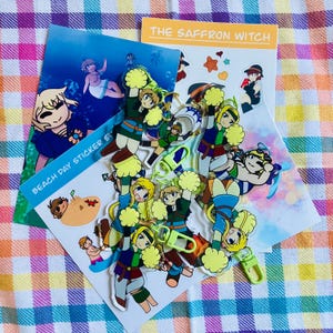 May include: A collection of stickers and keychains featuring anime-style characters. The stickers include the text "BEACH DAY STICKER SET" and "THE SAFFRON WITCH". The characters are depicted in various poses, some holding pom-poms. The background is a colorful plaid pattern.