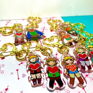 Può includere: Una collezione di portachiavi in acrilico trasparente con personaggi dei cartoni animati che tengono oggetti a forma di cuore rosso. Ogni portachiavi ha un fermaglio e un anello a forma di cuore color oro. I personaggi indossano vari abiti.