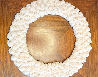May include: A cream-colored, circular wreath made of thick, looped yarn. The wreath is displayed on a wooden surface with a square pattern. The yarn has a soft, textured appearance, and the wreath's shape is clearly defined.