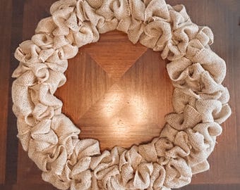 May include: A circular wreath made of beige burlap fabric. The burlap is gathered and ruffled, creating a textured, layered effect. The wreath is displayed on a dark brown wooden surface.