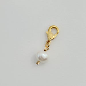 Op de afbeelding: Een goudkleurige bedel met een karabijnsluiting en een witte parel. De parel is rond en heeft een glad, glanzend oppervlak. De bedel is ontworpen voor het maken van sieraden of als accessoire.