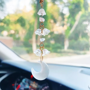 May include: A white crescent moon charm with a gold chain and white flower charms hanging from it. The charm is hanging from a rearview mirror in a car.