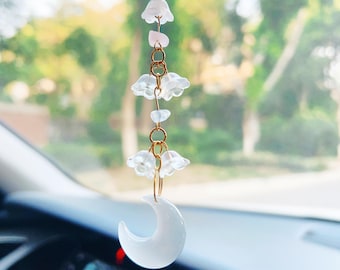 May include: A white crescent moon charm with a gold chain and white flower charms hanging from it. The charm is hanging from a rearview mirror in a car.