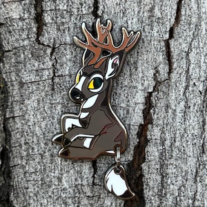May include: A silver enamel pin of a cartoon deer with brown and white stripes, large green eyes, and a brown antler. The deer is sitting with its tail dangling down. The tail is a separate piece of the pin and is white with a black tip.