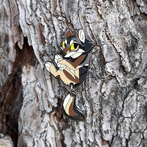 May include: A colorful enamel pin featuring a cartoon wolf with yellow eyes, in shades of black, brown, and tan. The wolf is hanging with its paws clasped. The pin has a dangling tail with brown, tan, and white stripes.