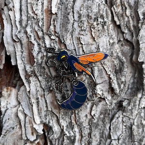 May include: A detailed enamel pin featuring a stylized insect with a blue body, orange wings, and a curved stinger. The pin includes a dangling, crescent-shaped element in dark blue and purple. The pin is set against a textured, gray and brown background.