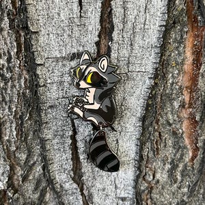 May include: A black and white enamel pin of a raccoon with yellow eyes, climbing a tree. The raccoon is holding a small object in its paws.