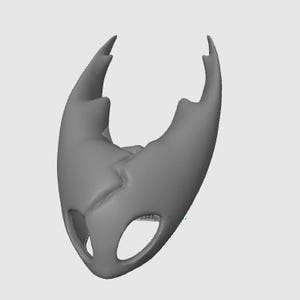 May include: A gray, stylized mask with a cracked design and two eye-shaped openings. The mask has two pointed protrusions at the top and a smooth, curved shape overall. The object is set against a plain, light gray background.