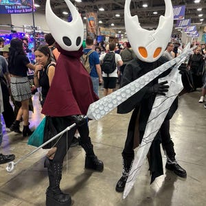 May include: Two cosplayers dressed as characters from the video game Hollow Knight. They wear white horned masks with black eye holes, capes, and black outfits. One holds a staff, the other a large, silver sword. The background shows a convention hall.