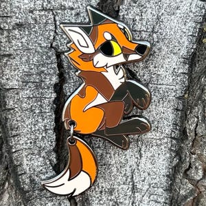 May include: A colorful enamel pin featuring a cartoon fox in shades of orange, brown, and white. The fox is in a sitting position with a silver outline. The pin has a small loop at the bottom for attachment.