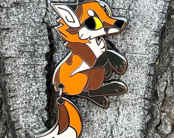 May include: A colorful enamel pin featuring a cartoon fox in shades of orange, brown, and white. The fox is in a sitting position with a silver outline. The pin has a small loop at the bottom for attachment.