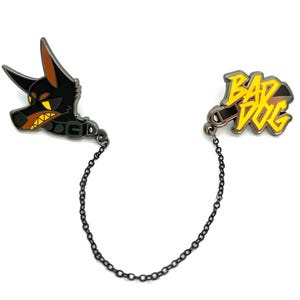 Op de afbeelding: Een zwart en bruin emaille pin set met een Dobermann hondenkop en de woorden "BAD DOG" in geel en zwart. De twee pins zijn verbonden door een zwarte ketting.