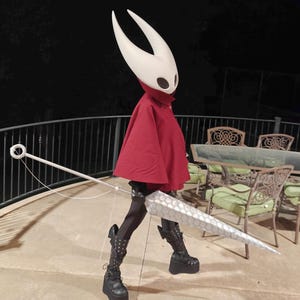 May include: A person in a cosplay costume featuring a white mask with two horns, a red cape, and a large, silver, geometric-patterned weapon. The person is wearing black platform boots and leggings. The background is a patio at night.