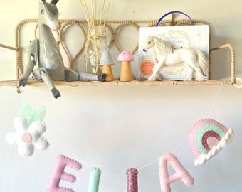 May include: A decorative shelf with various items, including a wooden deer figurine, a white toy horse, and felt mushroom decorations. A banner spelling "ELLA" hangs below, along with a felt flower and rainbow. The shelf is made of woven material.