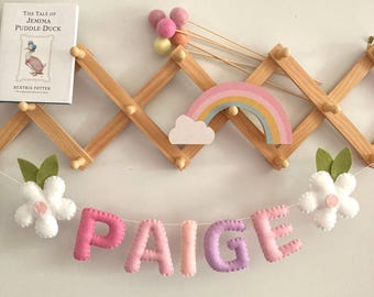 May include: A felt banner with the name "PAIGE" in pink and purple letters. The banner is strung with white felt flowers and green leaves. A rainbow and cloud are in the background.