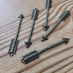 May include: Five antique gold arrow charms with a textured surface and a triangular arrowhead. The arrow charms have a decorative feathered tail.