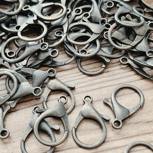 May include: A collection of antique bronze-toned metal lobster claw clasps. Each clasp features a spring-loaded mechanism and a small circular loop at the top for attachment. The finish is matte and slightly weathered, showing a dark metallic texture.