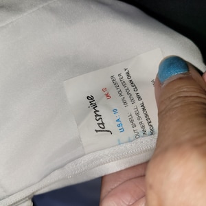 Może przedstawiać: Wnętrze białej odzieży z wszytą metką z nazwą marki Jasmine. Metka zawiera informacje: U.S.A.: 10, UK: 12, 100% poliester dla warstwy zewnętrznej i wewnętrznej oraz instrukcję dotyczącą wyłącznie profesjonalnego czyszczenia chemicznego.